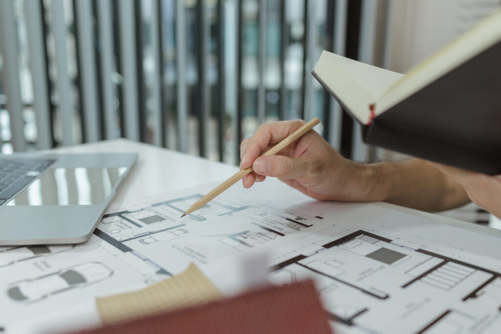 An architect reviewing detailed house floor plan drawings with a pencil, alongside a notebook and laptop on a work desk.