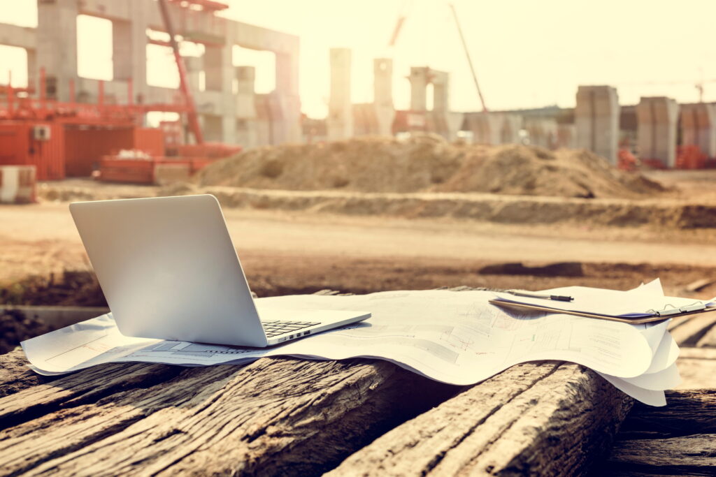 A laptop and construction drawings placed on a wooden surface at an active building construction site in Ontario, with structural frames and a crane visible in the background.