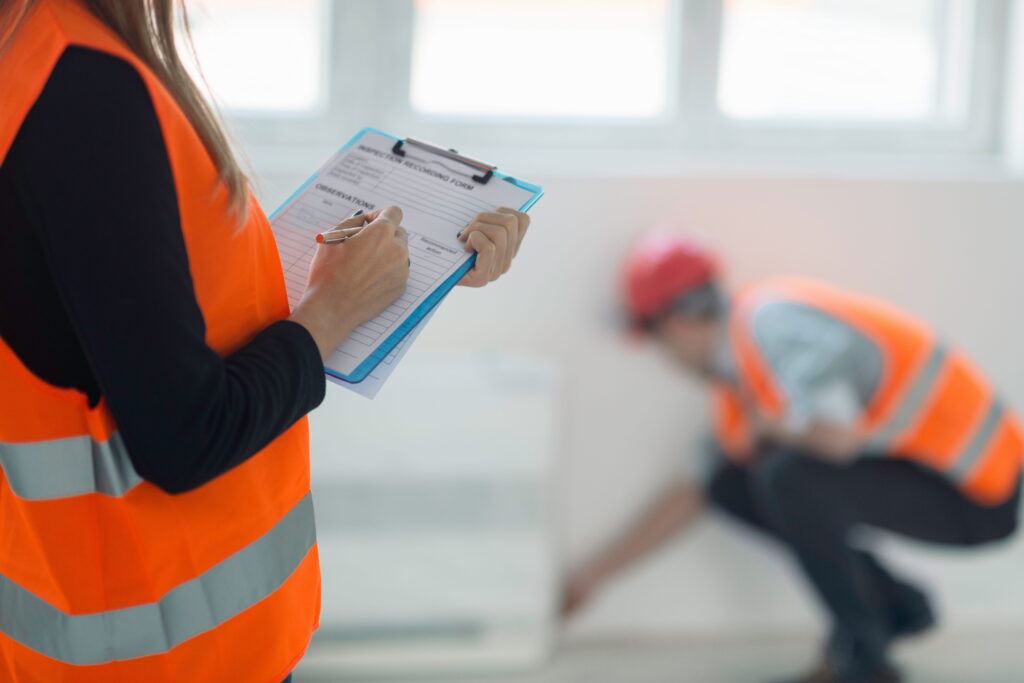 A home inspector in an orange safety vest completing an inspection recording form on a clipboard while a worker in a hard hat examines the wall in the background, representing the Tarion Pre-Delivery Inspection process for new homes in Ontario.